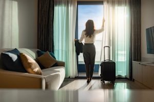 A woman opens her hotel blinds to reveal the ocean on a sunny day